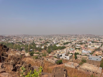 Vista aérea de la extensa ciudad de Bamako con edificios de colores variados bajo un cielo brumoso, con un primer plano de colinas rocosas y hierba seca