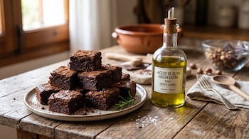 Brownies de chocolate en un plato cerámico junto a una botella con aceite de oliva y utensilios de cocina sobre una mesa de madera.