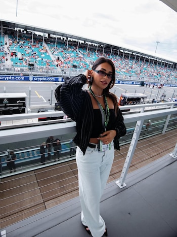 Mujer con gafas de sol, chaqueta oscura y pantalones claros posando en el borde de una pista de carreras con gradas llenas de gente al fondo