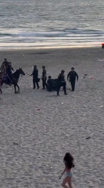 Una playa con agentes de policía a pie y a caballo, una persona en el suelo, una mujer en bañador y basura esparcida con el mar al fondo