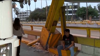 Parte frontal de un camión blanco, estructura metálica amarilla de puente con escombros en su base. Un hombre sentado, una mujer de espaldas. Fondo urbano
