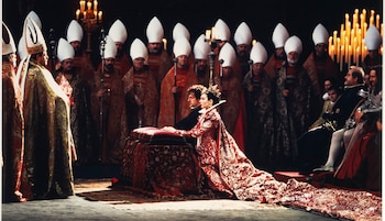 Una mujer vestida de rojo ornamentado y coronada se arrodilla frente a un hombre vestido de oro, rodeados por figuras con mitras blancas y público