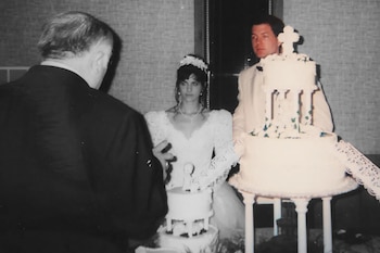 Una novia y un novio están de pie junto a un gran pastel de bodas blanco de varios niveles con una cruz arriba. Un hombre de espaldas en primer plano