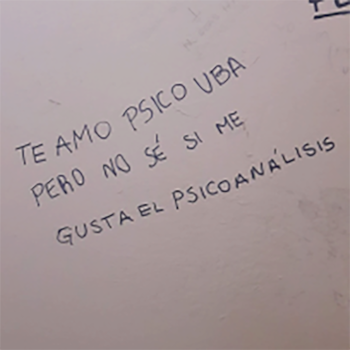 Primer plano de escritura a mano en tinta azul sobre una superficie blanca. El texto, en mayúsculas, dice 'TE AMO PSICO UBA PERO NO SÉ SI ME GUSTA EL PSICOANÁLISIS'