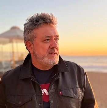 Retrato de medio cuerpo de un hombre con pelo gris y barba, vestido con chaqueta de cuero negra, mirando hacia la derecha con un atardecer sobre el mar de fondo