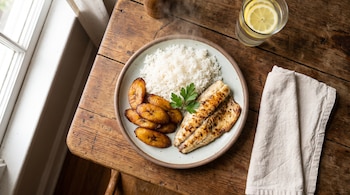 Plato con filete de pescado a la plancha, arroz blanco, plátanos fritos y perejil, acompañado por un vaso con agua y limón y una servilleta.