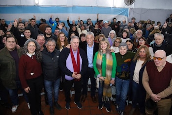 Un grupo grande de personas posa para una foto grupal en un salón con una bandera celeste y blanca al fondo. Varias personas levantan la mano derecha haciendo el signo de la victoria