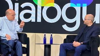 Dos hombres calvos sentados en sillones negros dialogan frente a una pantalla con la palabra 'Dialogu' y botellas azules sobre una mesa auxiliar