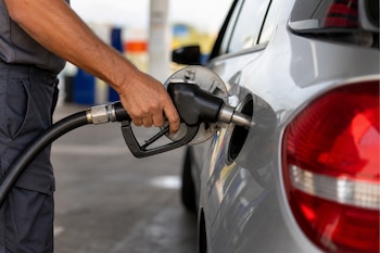 Plano detalle de una mano que sostiene la pistola de un surtidor para cargar combustible en el tanque de un automóvil gris.