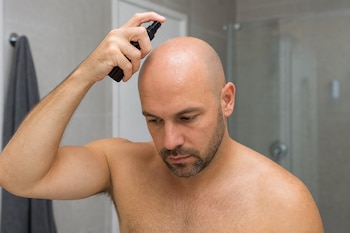 Hombre calvo y con barba que rocía un líquido desde un atomizador negro sobre su cabeza en un baño.