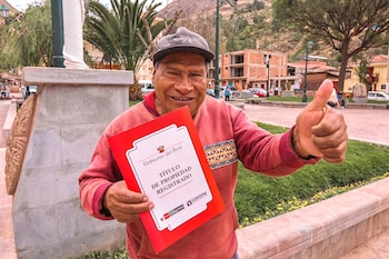 Un hombre con gorra y suéter rojo sostiene una carpeta roja de Cofopri y levanta el pulgar en una plaza frente a edificaciones y montañas