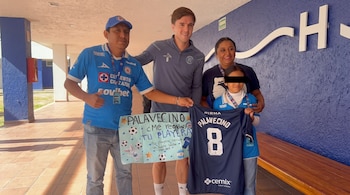 Un futbolista posa junto a dos adultos y una menor que sostienen una cartulina y una camiseta deportiva de color azul marino.