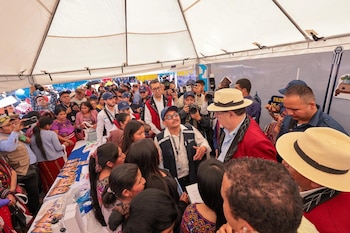 Gran grupo de personas diversas, algunas con sombreros de paja y otros sombreros, reunidas bajo una carpa blanca en un evento. Se ven cámaras y mesas con folletos