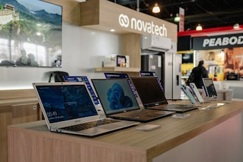 Tres computadoras portátiles, cuatro teléfonos inteligentes y una tableta en un mostrador de madera. Un letrero Novatech y electrodomésticos en segundo plano