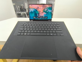 Portátil negro ASUS ROG Zephyrus Duo con pantalla principal encendida y teclado QWERTY en una mesa blanca. Se ve una mano sujetando el lateral derecho