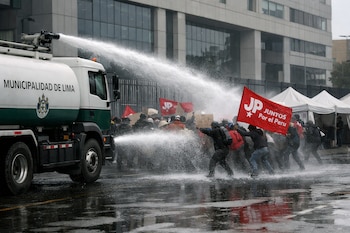 Camión cisterna de la Municipalidad de Lima rocía agua a manifestantes de Juntos Por el Perú con banderas rojas, en una calle mojada frente a un edificio institucional.