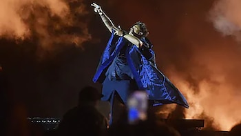 Un cantante vestido con una capa azul apunta hacia arriba en un escenario iluminado rodeado de humo