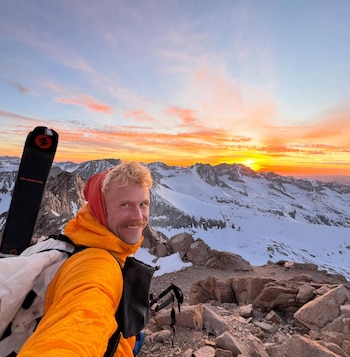 Jake Whisenant con chaqueta naranja y gorro rojo, esquís y bastones, en un pico rocoso nevado, con cielo de atardecer