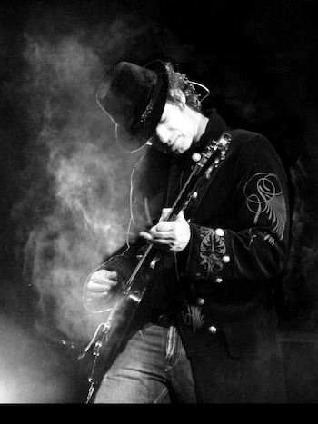 Imagen en blanco y negro de Gustavo Cerati tocando la guitarra eléctrica en un escenario con efectos de humo. Viste un sombrero de ala ancha y un abrigo oscuro decorado