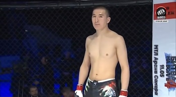 Joven luchador asiático sin camiseta en un octágono de MMA, con shorts negros estampados y guantes rojos y blancos. Hay una valla metálica de fondo y un cartel vertical a la derecha
