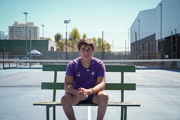 Un joven con camiseta morada y pantalones cortos oscuros se sienta en un banco verde en una cancha de tenis azul. La red y edificios se ven al fondo