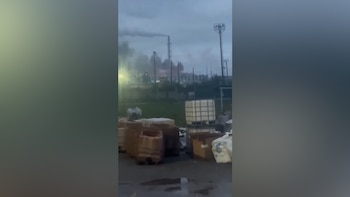 Contenedores y palés de madera en un patio exterior frente a una fábrica con chimeneas y humo bajo un cielo gris