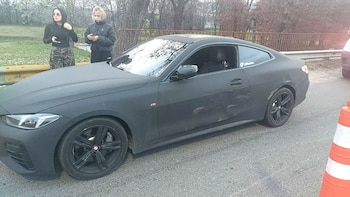 Un BMW deportivo negro mate con abolladuras en el lateral, estacionado en la calle. Dos mujeres jóvenes miran sus teléfonos detrás del vehículo