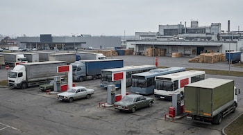 Múltiples camiones, autobuses y coches en una estación de servicio con surtidores rojos y grises. Al fondo, edificios industriales y un supermercado con palés.