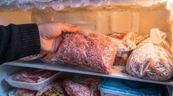 Primer plano de una mano colocando una bolsa transparente de carne picada cruda en un estante congelado. El congelador contiene otros paquetes de alimentos.