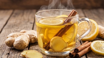 Primer plano de una taza de cristal con infusión humeante, rodajas de limón, jengibre y canela, sobre una mesa de madera rústica con ingredientes frescos.