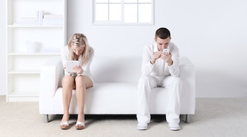 Un hombre y una mujer vestidos de blanco están sentados en un sofá blanco mientras miran una tableta y un teléfono móvil.
