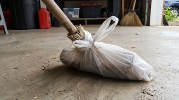 Una escoba con mango de madera y cerdas cubiertas por una bolsa de plástico blanca atada, sobre un suelo de cemento sucio dentro de un garaje.