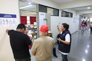 Varias personas, incluyendo votantes y personal de ONPE, en un pasillo de la Universidad Católica con un cartel de "Atención Preferencial" y número de mesa