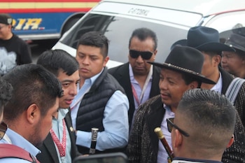 Varios hombres vestidos con sacos, sombreros y bastones de mando conversan cerca de una camioneta blanca y un autobús