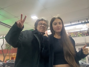 Un hombre con anteojos y cabello canoso rizado hace el gesto de la paz junto a una mujer joven de cabello largo