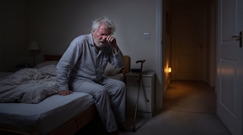 Hombre mayor con pijama de rayas sentado en la cama frotándose el ojo en la oscuridad. Hay un bastón y una luz tenue en el pasillo adyacente.