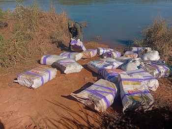 Un agente con uniforme verde levanta un bulto empaquetado junto a decenas de paquetes amontonados sobre la tierra cerca del agua