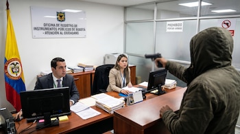 Una persona encapuchada empuña una pistola, apuntando a un hombre y una mujer sentados en escritorios de la Oficina de Registro de Instrumentos Públicos de Bogotá.