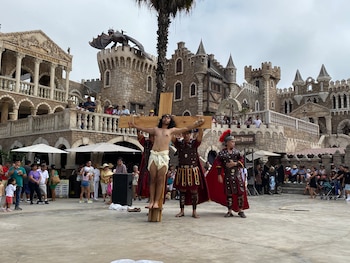 Representación teatral con un actor en una cruz de madera sostenida por otros dos vestidos de romanos, frente a un castillo temático con una estatua de dragón y público presente