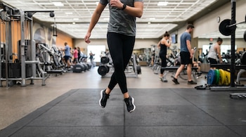 Persona con camiseta gris y leggings negros realizando un salto de skater en el aire dentro de un gimnasio. El suelo es de goma y hay equipos de ejercicio al fondo.