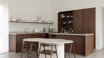 Cocina moderna con muebles de madera oscura, encimeras claras, fregaderos y electrodomésticos. Destaca una isla ovalada blanca con sillas de madera y estantes decorados.