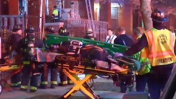 Un grupo de bomberos y paramédicos con uniformes y chalecos de alta visibilidad atiende a una persona en una camilla de ruedas en la calle, de noche