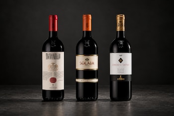 Tres botellas de vino tinto alineadas sobre una mesa oscura con cápsulas de color rojo, naranja y dorado