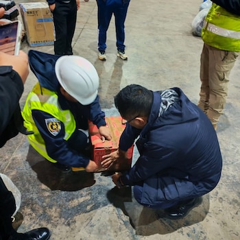 Dos hombres agachados examinan una caja de cartón en un piso de concreto, rodeados por otras personas con chalecos reflectantes