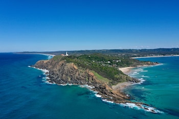 Vista aérea de un faro blanco sobre un promontorio rocoso y verde, rodeado por las aguas turquesas del océano y extensas playas de arena