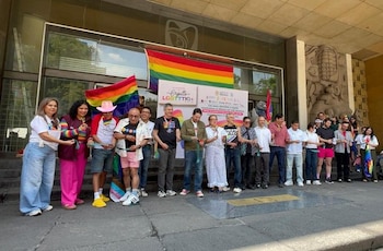 Un grupo de personas se encuentra frente a un edificio con una bandera arcoíris grande y un cartel. Varias personas sostienen banderas pequeñas de colores