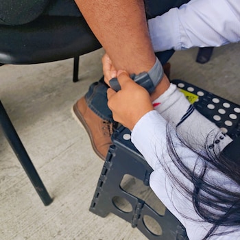 Primer plano de manos ajustando un dispositivo de monitoreo gris en el tobillo de una persona con pantalón de mezclilla y botas. Fondo: suelo de cemento
