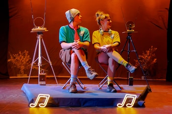 Dos personas jóvenes, hombre y mujer, sentadas en sillas de metal en un escenario oscuro con luces naranjas, frente a aparatos con antenas y notas musicales de neón