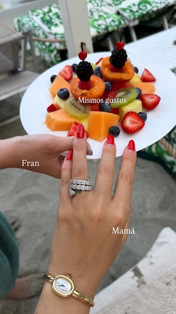 Dos manos, una con uñas rojas y un anillo, otra de una niña, sostienen un plato blanco con frutas picadas: melón, fresas, arándanos, kiwi, albaricoques y moras