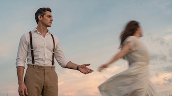 Un hombre de camisa blanca y tirantes extiende su mano hacia una mujer con vestido blanco en movimiento.